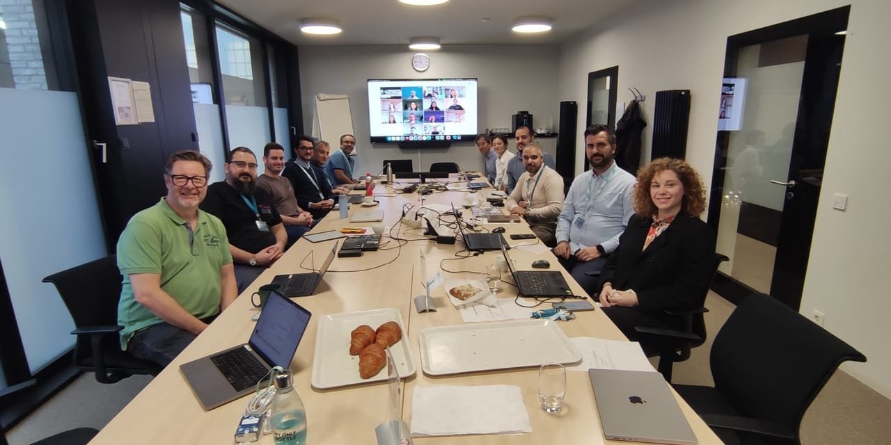 Kick-off meeting around a long conference table with laptops open, and remote partners shown on a video-call screen at the end of the room.