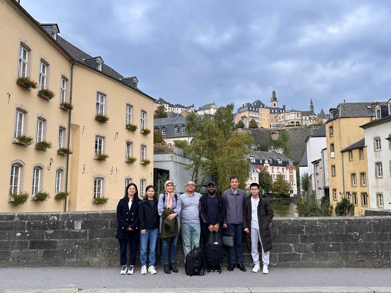 Seven members of the proposal team standing in a row on a stone bridge in Luxembourg City's Grund quarter, with yellow houses, the river and the Old Town on the cliffs behind them under an overcast sky.